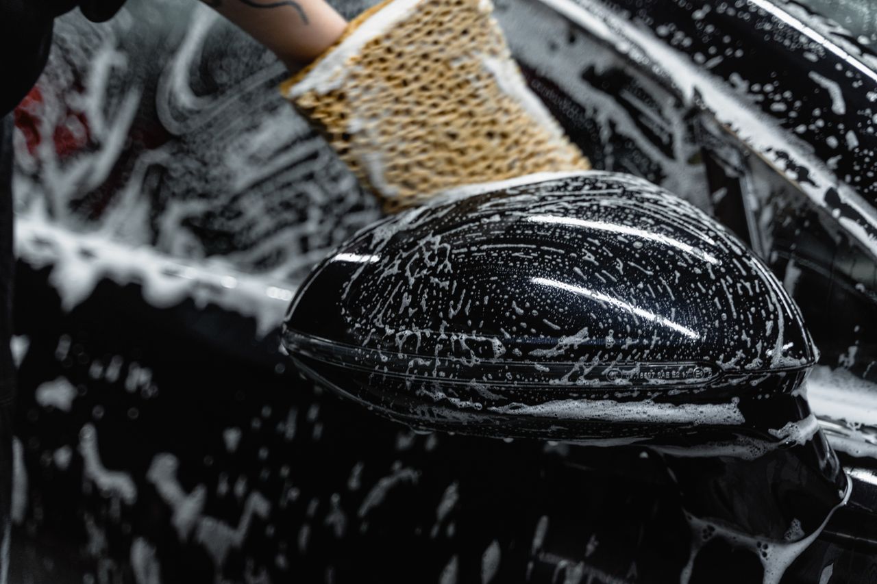 Hand scrubbing a car mirror with a sponge covered in soap at a car wash.